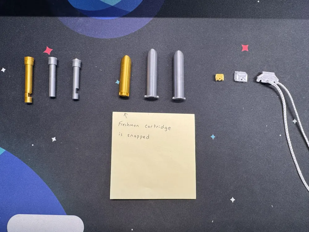Three groups of parts, freshman versions in gold beside the grey rebuilds: bullets, cartridges and stoppers. A sticky note reads: Freshman cartridge is snapped.