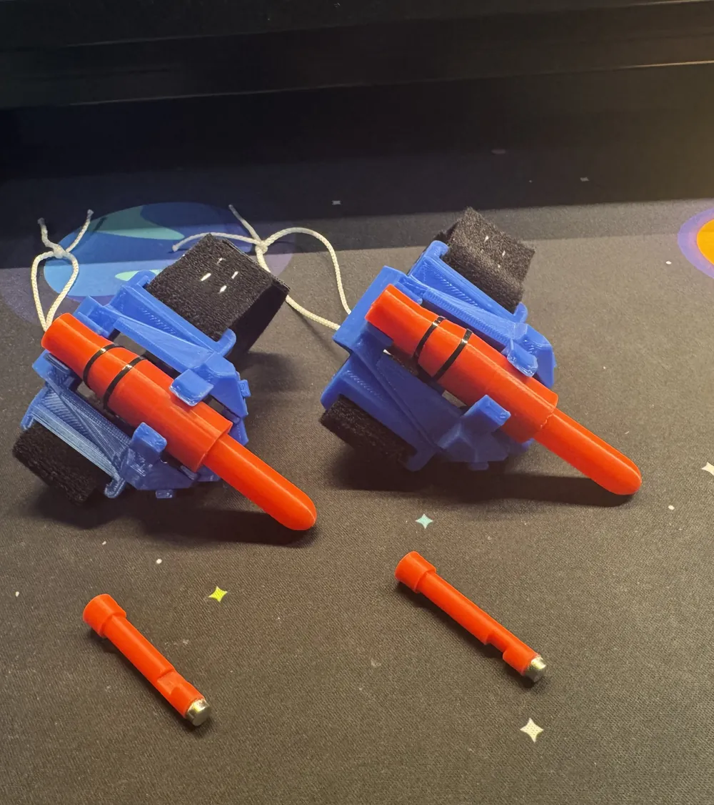 Two finished webshooters side by side: blue casings, red barrels, black Velcro straps and white pull strings, with two spare red bullets in front showing the magnets on their tips.