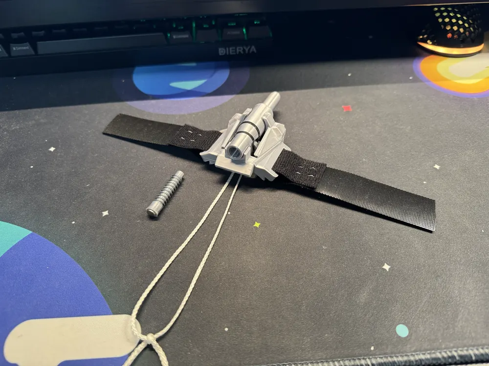The assembled V2 webshooter on its black Velcro strap, lying on a desk mat beside a spare bullet and spring, pull strings trailing.