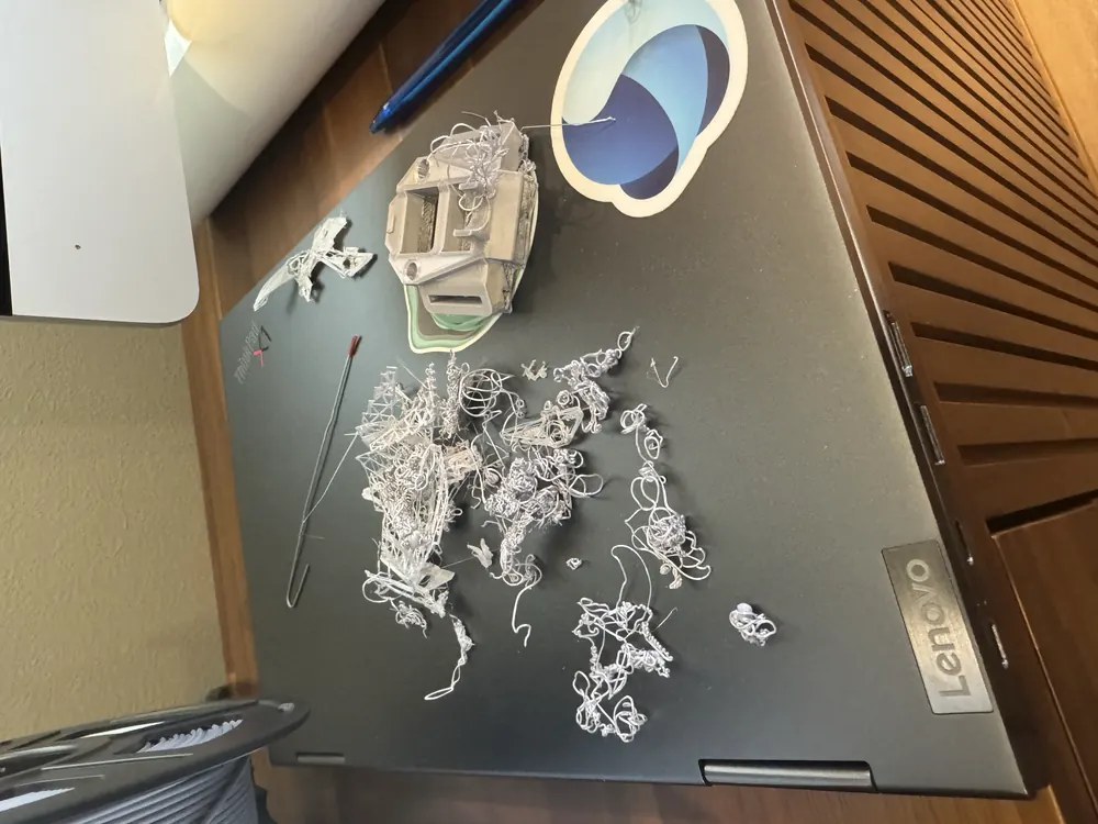 A tangled heap of failed grey filament spaghetti next to a casing part on a laptop.